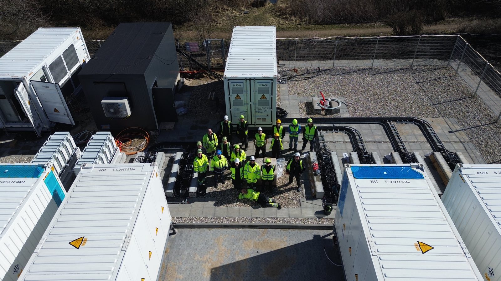 Medarbejdere i sikkerhedsveste og hjelme samlet mellem battericontainere på et anlæg under opførelse.
