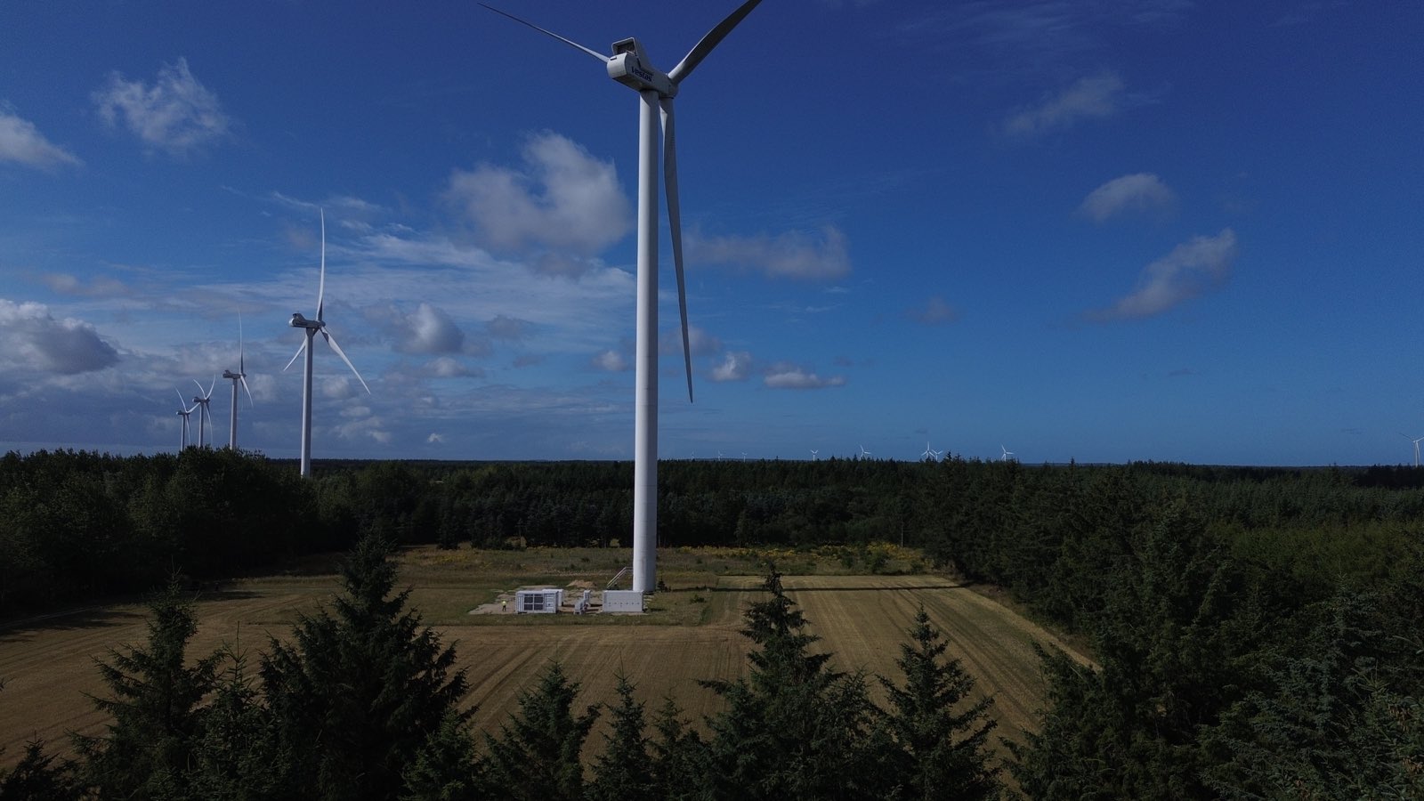 Luftfoto af en co-location-park: et batterilagringsanlæg placeret ved foden af en vindmølle i et skovområde, med flere møller i baggrunden.